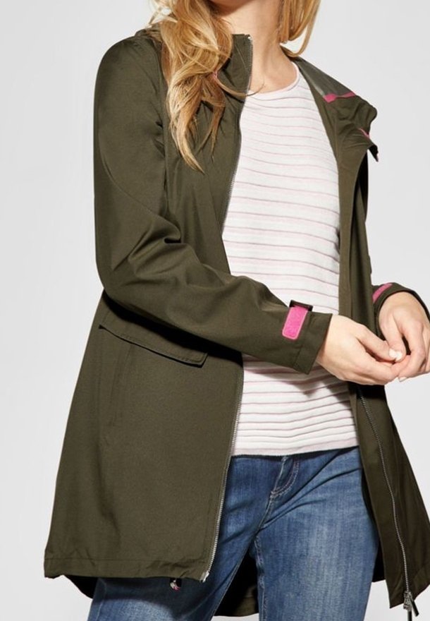 Femme portant une veste à capuche vert olive avec des détails roses sur une chemise blanche rayée et un jean bleu, ajustant sa manche.