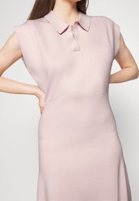 Femme portant une robe en maille rose clair sans manches avec col, debout les mains sur les hanches devant un fond uni.