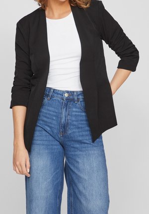Kvinde iført en sort blazer, hvid skjorte og blå taljehøje jeans med venstre hånd hvilende på hoften.