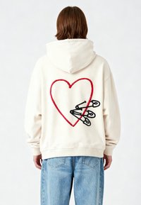 Persona che indossa una felpa bianca con una grafica di un cuore rosso e spilli di sicurezza neri sulla schiena, abbinata a jeans azzurri, di spalle.