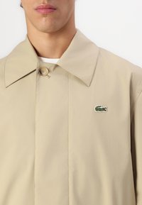 Beige letvægtsjakke med en stor krave, enkel knaplukning og et grønt krokodillelogo broderet på venstre side.