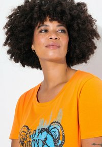 Orange T-shirt i blødt bomuld med et blåt grafisk design med prikker og linjer. Korte ærmer, rundt halsudskæring og en afslappet pasform.