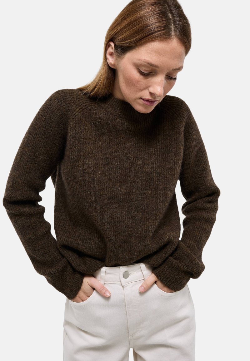 Femme aux cheveux bruns raides portant un pull en tricot marron foncé et un pantalon beige clair, les mains dans les poches, regardant vers le bas.