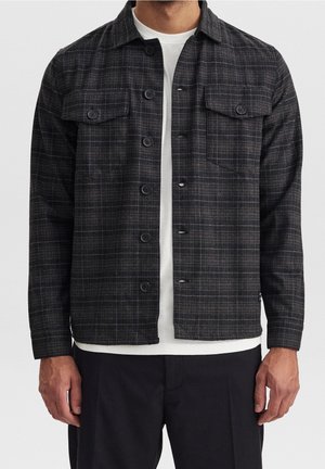 Homme portant une chemise-veste à carreaux noire et grise boutonnée par-dessus un t-shirt blanc et un pantalon noir, debout les bras détendus.