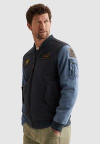 Hombre con una chaqueta bomber azul marino y de mezclilla con parches, mirando hacia un lado con las manos en los bolsillos de pantalones caqui.