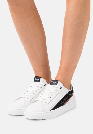 Pieds chaussés de baskets basses blanches avec des accents noirs et or rose et des lacets blancs sur un fond clair uni.