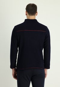 Marineblauwe fleece pullover met een kraag, voorzien van contrasterende rode stiksels langs de schoudernaad. Lange mouwen en rechte zoom. Zachte textuur.