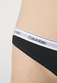 Czarne majtki Calvin Klein z szarym elastycznym pasem w logo. Gładki materiał, niski krój oraz obszyte krawędzie dla zwiększonej trwałości.