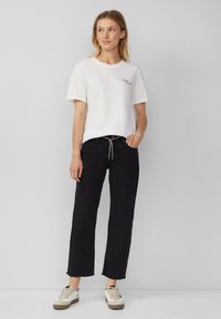 T-shirt en coton blanc avec texte brodé rouge, associé à un pantalon large noir et des baskets beiges et blanches ; coupe et design décontractés.