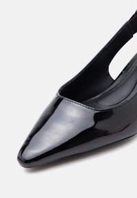 Μαύρο γυαλιστερό δερμάτινο παπούτσι με μυτερό δάχτυλο και λουρί πίσω, χαμηλό τακούνι και λεία επιφάνεια, σε φωτεινό φόντο.