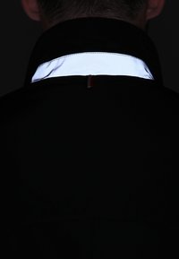 Černá bunda s reflexním bílým límcem a červeným detailem na zádech, která v nízkém osvětlení odhaluje hladkou texturu.