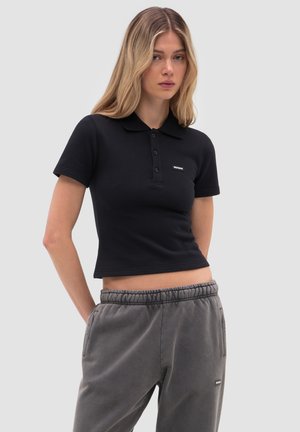 Młoda kobieta z długimi blond włosami, ubrana w dopasowaną czarną koszulkę polo i szare spodnie dresowe z elastycznym pasem, na tle jasnego, jednolitego tła.