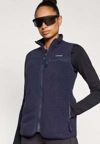 Μπλε ναυτικό γιλέκο από φλις με ψηλό γιακά, φερμουάρ στο μπροστά μέρος και δύο πλαϊνές τσέπες. Διαθέτει υφή υφάσματος και λογότυπο στο στήθος.