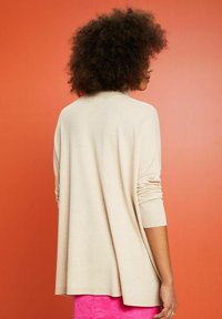 Beżowy, oversize sweter z prążkowanymi mankietami, o luźnym kroju i miękkiej fakturze. Tło w gradientowym pomarańczowym kolorze.