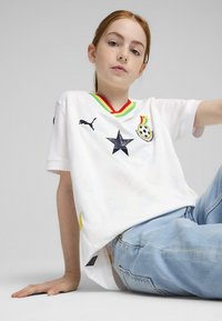 Balts futbola kreklis ar melnu zvaigznes emblem, varavīksnes apdari un Ganas karoga akcentiem. Apvienots ar gaiši ziliem džinsiem.