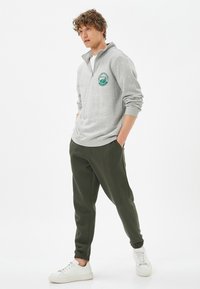 Jasnoszary sweter z zamkiem przy kołnierzu i zielonym logo, w zestawieniu z ciemnozielonymi spodniami dresowymi i białymi sneakersami. Miękka tkanina, luźny krój.