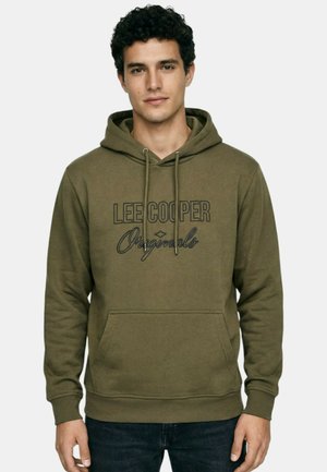 Homme aux cheveux bouclés portant un sweat à capuche vert olive avec le texte "Lee Cooper Originals" et un jean foncé, debout devant un fond uni.