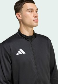 Μαύρο αθλητικό μπουφάν με φερμουάρ μπροστά, ψηλό γιακά και τρία λευκά λογότυπα στη πάνω αριστερή πλευρά του στήθους. Λείος υφασμάτινος χαρακτήρας και εφαρμοστό σχέδιο.