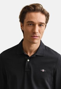 Homme aux cheveux châtain clair, aux yeux bleus, portant un polo noir à col avec un petit logo sur la poitrine, expression neutre.