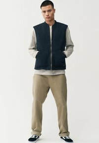 Marineblauw mouwloos vest met een ritssluiting aan de voorkant en twee zakken, gedragen over een grijs-wit gestreept langetermijnshirt, in combinatie met beige broeken.
