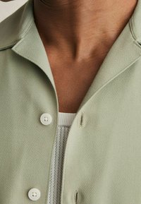 Camisa abotonada de espiga verde claro con cuello, que presenta botones blancos y una camiseta interior blanca acanalada asomando por el escote.