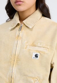 Veste en denim beige dotée d'un col en cordon, d'une fermeture éclair et d'une poche plaquée. Un petit logo noir et blanc Carhartt est affiché sur la gauche.
