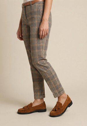 Osoba nosící béžové, námořnicky modré a oranžové kárované kalhoty s hnědým opaskem a hnědými semišovými mokasíny s třásněmi na jednobarevném pozadí.