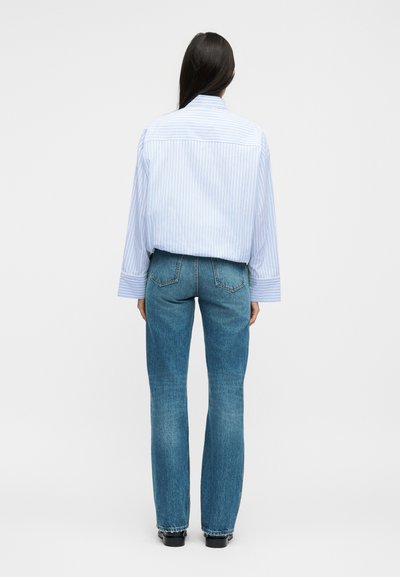 Γυναίκα με μακριά σκούρα μαλλιά, που στέκεται με την πλάτη, φοράει ανοιχτό μπλε πουκάμισο με ρίγες και φαρδιά μανίκια και μπλε τζιν κανονικής γραμμής.