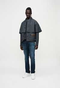 Σύντομο γκρι αδιάβροχο μπουφάν με φαρδιά μανίκια, κίτρινο φερμουάρ ως διακριτικό στοιχείο, σε συνδυασμό με μπλε τζιν και λευκά αθλητικά παπούτσια. Ομαλή υφή.