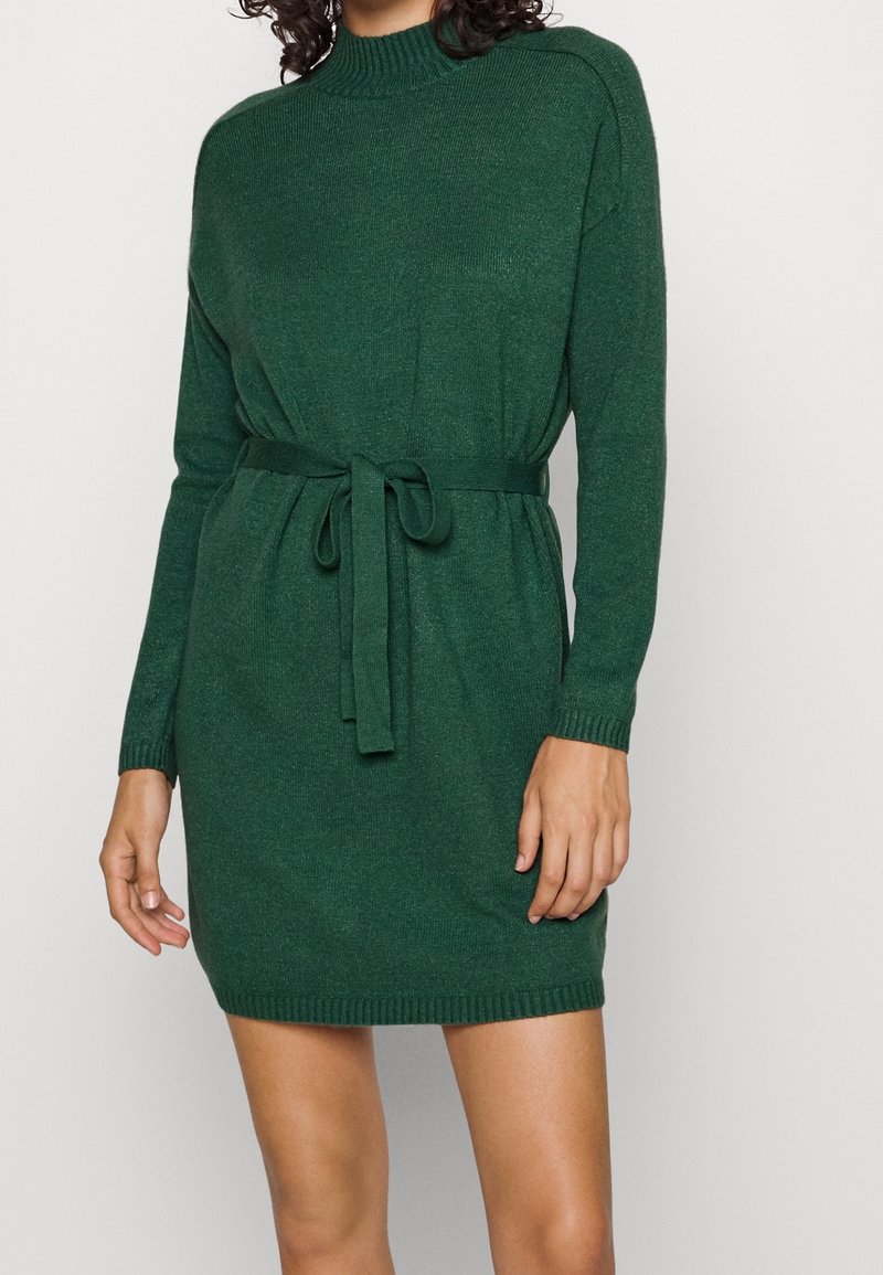 Femme portant une robe en tricot vert foncé à manches longues, avec un col montant et une ceinture assortie nouée à la taille.