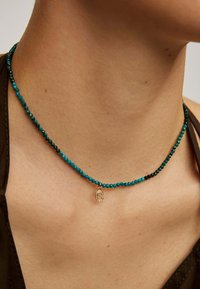 Collar de cuentas con colores turquesa, negro y dorado; incluye un pequeño encanto dorado en forma de la letra 'C'.