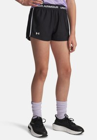 Person klädd i svarta Under Armour träningsshorts, lila scrunched strumpor, svarta löparskor och en lila topp, stående med ena handen på höften.