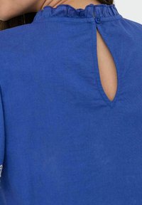 Top in tessuto blu con colletto arricciato, chiusura con bottoni sulla schiena e dettaglio a apertura a chiave. Texture liscia e design casual.