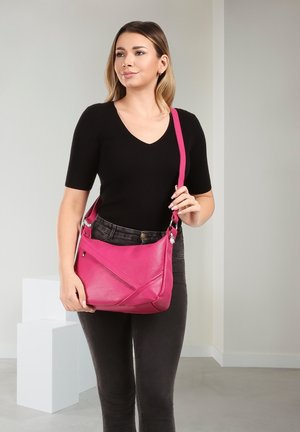 Femme en haut noir et jean tenant un sac à bandoulière en cuir rose vif, debout à l'intérieur sur un fond neutre.