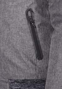 Grå texturerad tyg med ett fiskbensmönster, med en svart dragkedjeficka på vänster sida och mörkgrå stickade detaljer i botten.