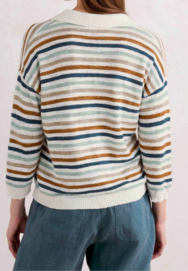 Femme portant un pull en maille blanc avec des rayures horizontales marron, vertes et bleues, associé à un pantalon bleu ample, vue de dos.