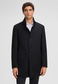 Cappotto di lana nera con colletto alto, chiusura frontale a bottoni e tasche laterali. Texture liscia e silhouette sagomata, adatto per abbigliamento formale.