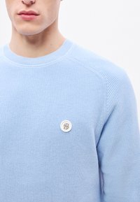 Világoskék kötött pulóver bordázott mintával. Kerek nyakkivágással és ezüst színű logógombbal a bal mellrészen.