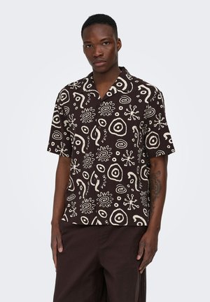 Homme portant une chemise noire à manches courtes avec des motifs abstraits blancs en spirale et soleil, associée à un pantalon foncé, debout devant un fond uni.