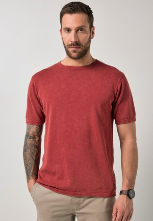 Hombre con barba que lleva una camisa roja de manga corta, pantalones beige, brazo izquierdo tatuado y un reloj en la muñeca derecha, posando contra un fondo liso.