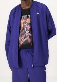 Lila lättviktig jacka med dragkedja, elastiska muddar och Adidas-logotyp. Svart grafisk t-shirt under med livfulla sportbilder och text.