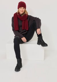 Črne vezalne gležnjarje, temne kavbojke, sivi rebrasti pulover ter maroon pleteni šal in kapa, sedi na belem kubusu proti svetli ozadju.