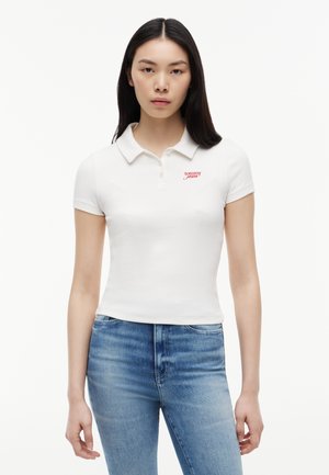 Naine kannab valget lühikeste varrukatega polo särki, millel on "tommy jeans" logo, ja siniseid teksaseid, seisab ühevärvilise valge tausta ees.