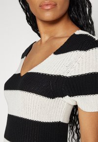 Schwarz-weißer gestreifter Strickoberteil mit V-Ausschnitt, kurzen Ärmeln und texturiertem Garn, das abwechselnde Farbmustern zeigt.