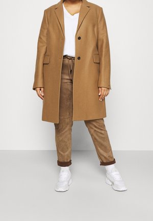 Persoon draagt een camelkleurige jas, wit V-hals T-shirt, bruine suède broek en witte sportschoenen, staand tegen een effen achtergrond.