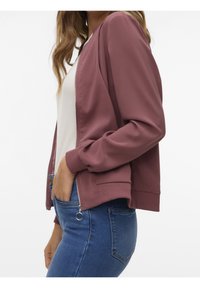 Bordeauxrode bomberjack met een rits aan de voorkant, geribbelde manchetten en zoom. Lichtgewicht stof met een gladde textuur. Gedragen over een wit shirt en blauwe spijkerbroek.