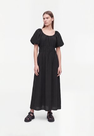 Vrouw staand in een zwarte midi-jurk met pofmouwen, een geplooide taille en zwarte sandalen met gesloten neus tegen een effen witte achtergrond.