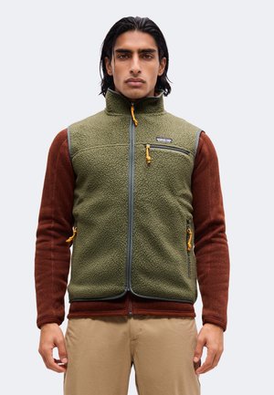 Homme portant une veste polaire vert olive par-dessus une chemise à manches longues couleur rouille et un pantalon beige sur fond uni.