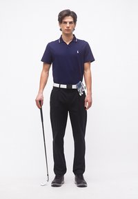 Νεαρός άνδρας στέκεται κρατώντας μπαστούνι γκολφ, φορώντας μπλε πόλο, μαύρο παντελόνι, λευκή ζώνη και γάντι γκολφ που είναι προσαρτημένο στη ζώνη.