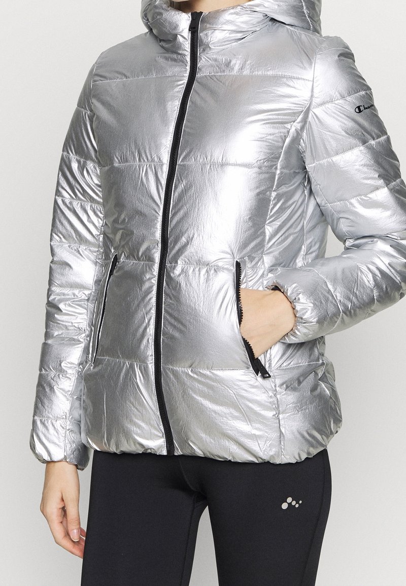 Femme portant une doudoune argentée brillante avec capuche, fermeture éclair noire et poches zippées, associée à un legging noir.